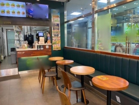"안양 프랭크버거 인수 매장의 홀 인테리어 전경. 쾌적한 녹색 소파 좌석과 원목 테이블, 그리고 뒤로 보이는 청결한 오픈 주방과 키오스크 주문 공간"