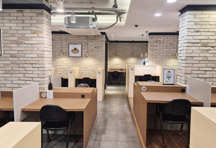 화이트 벽돌 기둥과 따뜻한 원목 소재의 칸막이 책상, 검은색 의자가 아늑하게 배치된 프리미엄 무인 스터디카페 내부 인테리어. 무인 시스템으로 운영 가능한 유망 1인 창업 아이템 예시 비주얼.