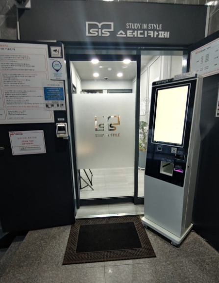 평촌독서실 양도양수 매물로 나온 SIS스터디카페 안양평촌1호점의 출입구 전경입니다. 무인 출입 관리를 위한 키오스크와 깔끔하게 정돈된 입구 시스템이 잘 갖추어져 있습니다.